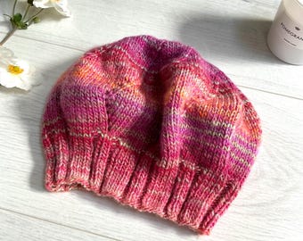 Könnte beinhalten: Handgestrickte Beanie-Mütze in Rosa-, Orange- und Cremetönen. Die Mütze hat einen gerippten Bund und ein strukturiertes Strickmuster. Die Mütze ist aus weichem, warmem Garn gefertigt, ideal für kaltes Wetter.