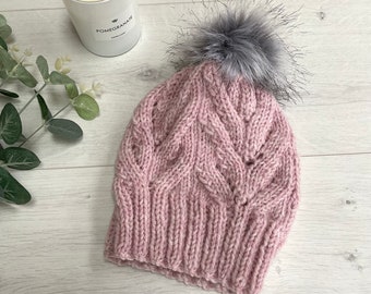 Könnte beinhalten: Ein gestrickter rosa Beanie mit einem grauen Kunstfell-Bommel. Die Mütze hat ein Zopfmuster und einen gerippten Saum. Die Mütze ist aus einem weichen, warmen Material gefertigt. Die Mütze ist etwa 23 cm hoch.