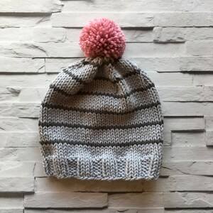 Puede incluir: Un gorro de punto con rayas horizontales en gris claro y gris oscuro, acabado con un borde acanalado en la parte inferior y un pomp&oacute;n rosa grande y texturizado unido a la corona del sombrero, creando un aspecto suave y c&aacute;lido.