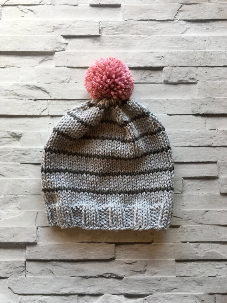 Puede incluir: Un gorro de punto con rayas horizontales en gris claro y gris oscuro, acabado con un borde acanalado en la parte inferior y un pomp&oacute;n rosa grande y texturizado unido a la corona del sombrero, creando un aspecto suave y c&aacute;lido.
