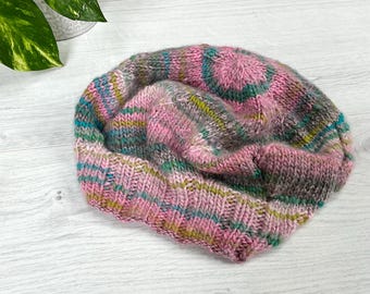 Könnte beinhalten: Eine handgestrickte Beanie-Mütze aus einer Mischung aus rosa, türkis, grünen und grauen Garnen. Die Mütze hat einen gerippten Saum und eine strukturierte, mehrfarbige Krone. Die Mütze ist aus weichem, warmem Garn gefertigt.