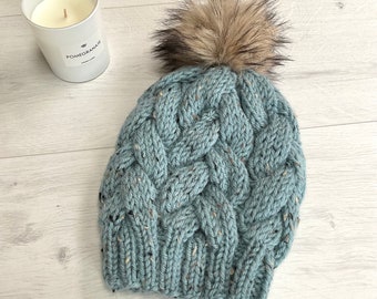 Könnte beinhalten: Hellblaue Zopfmuster-Beanie mit einem Kunstfell-Bommel. Die Mütze hat ein strukturiertes, gewebtes Design und Farbtupfer. Eine weiße Kerze mit dem Wort "Pomegranate" ist im Hintergrund zu sehen.