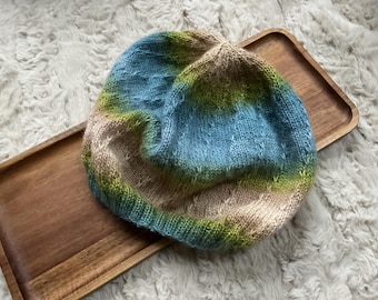 Könnte beinhalten: Handgestrickte Beanie-Mütze mit Farbverlauf in Blau, Grün und Beige. Die Mütze liegt auf einem Holztablett vor einem flauschigen, weißen Hintergrund.