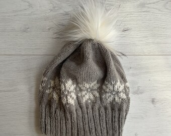Könnte beinhalten: Graue Strickmütze mit weißem Schneeflockenmuster und einem großen weißen Kunstfell-Bommel. Die Mütze ist aus weichem, strukturiertem Garn gefertigt, ideal für kalte Tage.