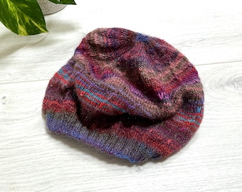 Könnte beinhalten: Handgestrickte Beanie-Mütze in einer Mischung aus Burgunderrot, Lila und Blau. Die Mütze hat einen gerippten Bund und eine strukturierte, mehrfarbige Krone. Das Garn scheint einen leichten Glanz zu haben, was ihren visuellen Reiz erhöht.