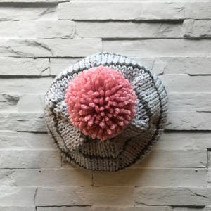 Puede incluir: Un gorro de punto con una base de hilo gris claro, rayas verticales gris oscuro y un pomp&oacute;n rosa grande y texturizado en la parte superior central. El gorro est&aacute; confeccionado con un patr&oacute;n de punto acanalado en toda la superficie exterior.