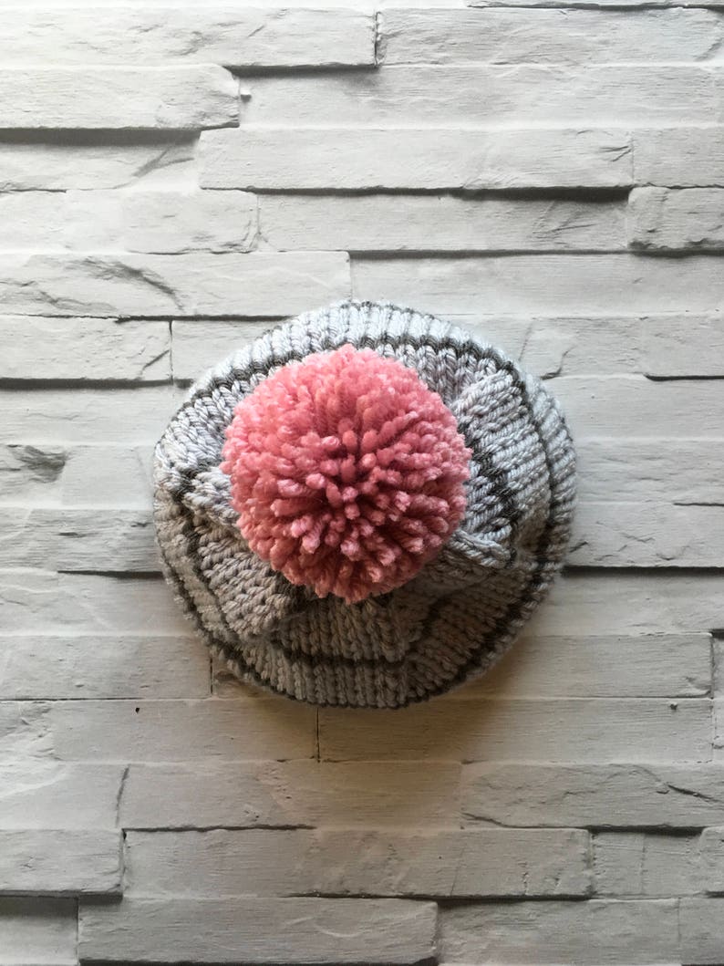 Puede incluir: Un gorro de punto con una base de hilo gris claro, rayas verticales gris oscuro y un pomp&oacute;n rosa grande y texturizado en la parte superior central. El gorro est&aacute; confeccionado con un patr&oacute;n de punto acanalado en toda la superficie exterior.