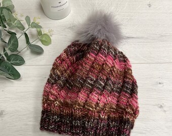 Könnte beinhalten: Eine handgestrickte Wintermütze mit einem grauen Kunstfell-Bommel. Die Mütze hat ein strukturiertes Muster in Rosa-, Braun- und Cremetönen. Ein warmer und stilvoller Winter-Accessoire.