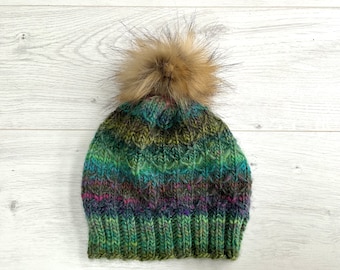 Könnte beinhalten: Handgestrickte Beanie-Mütze mit strukturiertem Muster in Grün-, Blau- und Lilatönen. Die Mütze ist mit einem großen, flauschigen, braunen Kunstfell-Bommel versehen. Die Mütze liegt auf einer hellen Holzmaserung.