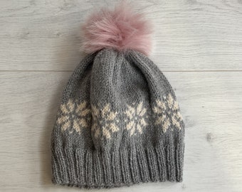 Könnte beinhalten: Graue Strickmütze mit einem hellrosa Kunstfell-Bommel. Die Mütze hat ein Schneeflockenmuster in Creme. Die Mütze ist aus einem weichen, warmen Material gefertigt, ideal für kaltes Wetter.
