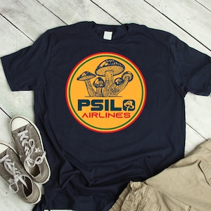 Op de afbeelding: Marineblauw T-shirt met een cirkelvormig ontwerp van paddenstoelen en de tekst "PSILO AIRLINES". Het shirt wordt gecombineerd met grijze sneakers en een kaki broek.