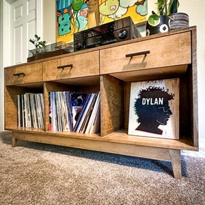 May include: A wooden record player stand with two drawers and three shelves. The shelves are filled with vinyl records. A framed print with the word "DYLAN" is on the top shelf.