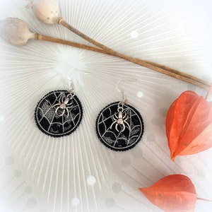 Peut inclure: Une paire de boucles d'oreilles en forme de toile d'araignée noire avec des breloques d'araignée argentées. Les boucles d'oreilles sont sur un fond blanc avec des fleurs séchées.