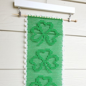May include: A green crocheted wall hanging with a white crocheted border. The hanging is attached to a white wooden dowel rod with metal clips.