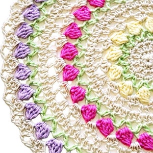 Könnte beinhalten: Nahaufnahme einer handgefertigten Häkeldecke. Die Decke hat eine cremefarbene Basis mit Reihen von lila, rosa und gelben floralen Akzenten. Das offene Design ist über das gesamte Stück sichtbar und erzeugt ein zartes und strukturiertes Aussehen.