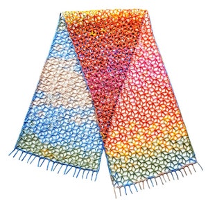 Op de afbeelding: Een kleurrijke gehaakte sjaal met een geometrisch patroon en franjes. De sjaal is gemaakt in tinten blauw, groen, geel, oranje, roze en rood.