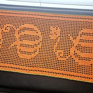May include: An orange crochet table runner featuring the word "BOO". The handmade runner has a decorative edge and is ideal for Halloween. The orange colour is vibrant and the open weave design is visible.