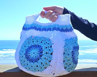 May include: White crochet tote bag with blue and light blue granny squares. The bag has a single handle and is held by a hand against a blue sky and ocean background.