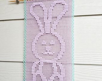 Op de afbeelding: Een lavendelkleurige gehaakte wandhanger met een delicaat bloemenmotief, omlijst door een lichtblauwe gehaakte rand. De hanger wordt getoond op een witte houten muur.