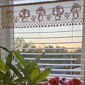 Peut inclure: Un lambrequin de fenêtre beige et crocheté avec un motif floral répété est suspendu au-dessus d'une fenêtre avec des stores blancs. La cantonnière est bordée d'une bordure festonnée. En dessous, un arrangement floral vibrant et une plante verte feuillue sont visibles.