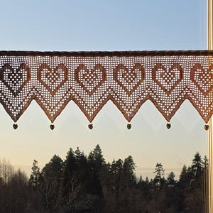 May include: A brown, crocheted window valance with a heart pattern. The valance is suspended in front of a window, with a forest visible in the background. The valance has a scalloped edge with heart-shaped pendants.
