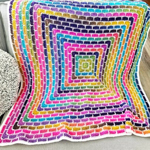 Può includere: Una coperta all'uncinetto colorata con un motivo quadrato in tonalità di rosa, viola, blu, verde, giallo e arancione. La coperta è realizzata con un filo bianco e ha un bordo bianco.