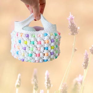 May include: A white crocheted basket with a handle, decorated with colorful beads. The basket is filled with white and pastel-colored balls.