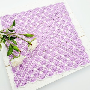 May include: A delicate lavender crochet doily with a white border. The doily is laid on a white wooden surface with a sprig of white flowers.