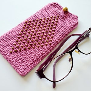 May include: A pink crocheted glasses case with gold beads and a gold button closure. The case is lying next to a pair of purple-framed glasses.