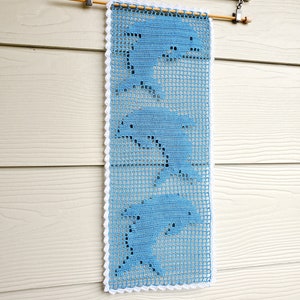 May include: A light blue crochet wall hanging with three dolphin designs. The rectangular piece has a white scalloped border and is suspended from a wooden dowel rod. The wall hanging is displayed against a light-coloured wooden background.