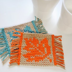 May include: Two crocheted coasters with fringe. One coaster is beige with orange geometric shapes and the other is beige with turquoise geometric shapes.