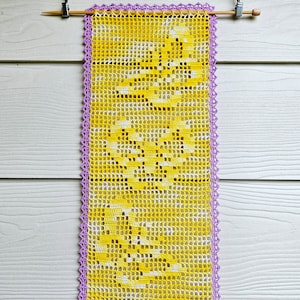 May include: A yellow and white crocheted wall hanging with a purple crocheted border. The hanging is rectangular and is attached to a white wooden dowel rod with two silver clips.