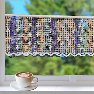 Peut inclure: Une cantonnière de fenêtre crochetée avec un motif floral dans les tons de violet, pêche et bleu. La cantonnière est suspendue devant une fenêtre avec un cadre blanc. Une tasse de café blanche avec du latte art est posée sur le rebord de la fenêtre.