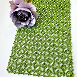 Op de afbeelding: Groene gehaakte tafelloper met een ruitpatroon. De loper ligt op een wit oppervlak met een paarse roos.