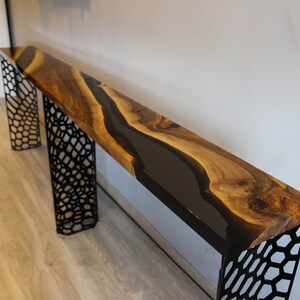 Può includere: Una lunga consolle in legno con un fiume di resina epossidica scura e lucida al centro. Il legno ha una venatura naturale e toni caldi. Il tavolo è sostenuto da due gambe in metallo nero geometriche, lungo circa 200 cm.