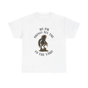 Peut inclure: T-shirt blanc avec un motif graphique représentant une créature et le texte "MY DM BRINGS ALL THE TO THE YARD" disposé en cercle. Le motif est dans les tons marron et noir.