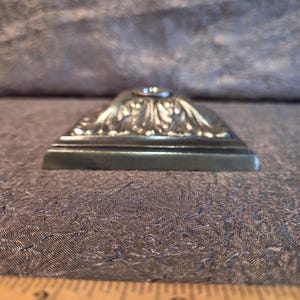May include: A silver-toned, rectangular decorative object with ornate floral detailing on the top surface. The object has a small, circular indentation at the center and sits on a textured surface.