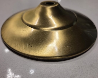 May include: A brass-colored, decorative metal base with a circular, slightly concave design. The base has a central hole and a smooth, polished finish, reflecting light. The overall shape is reminiscent of a small lampshade or a decorative element.