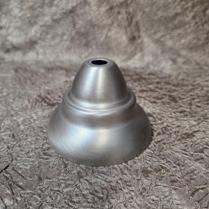 May include: A brushed nickel ceiling canopy with a smooth, tiered, bell-shaped profile. The metal component features a circular opening at the top centre and a wide, flared base, designed for mounting light fixtures to a ceiling surface.