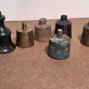 May include: A collection of antique metal bell-shaped objects in various sizes and finishes. The objects range in color from bronze to silver, with some showing signs of age and patina. The objects are arranged on a brown surface.