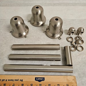 May include: A set of silver-toned metal light fixture parts, including three lamp holders, three metal rods, two finials, and four metal rings. The parts are arranged on a white surface with a wooden ruler labeled "Lowe's" in the bottom right corner.