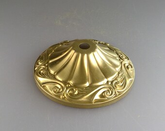 May include: A gold metal lamp finial with a decorative, scalloped design and a central hole. The finial has a detailed, swirling pattern around the edge.