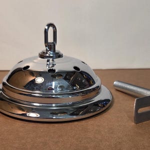 May include: A polished chrome ceiling fan canopy with a hanging loop and small ventilation holes. A matching metal bracket and threaded rod are next to the canopy. The items are on a brown surface.