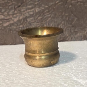 May include: A small, antique brass mortar with a flared rim and rounded base. The mortar has a warm, golden hue and shows signs of age and use. It is likely used for grinding herbs or spices.