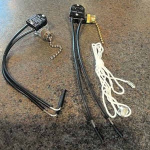 May include: Two black electrical light switches with attached wires. One has a pull chain and a brass fitting. The other has a small glass bulb and a metal chain. Both have black plastic housings.