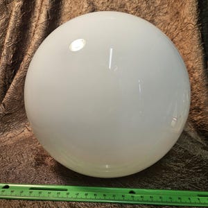 以下が含まれることがあります： 大きな不透明な白い球体、おそらくガラスまたはプラスチックが画像全体を占めています。緑色の定規が球体の下に配置され、サイズの目安となっています。球体の表面は光を反射しています。背景は質感のある茶色の布です。
