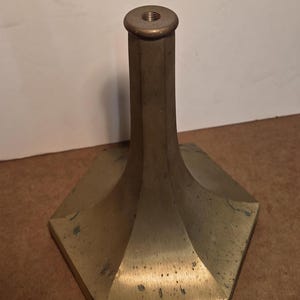 May include: A brass-colored metal candlestick base with a hexagonal base and a cylindrical top. The base has a flared design, and the top has a threaded opening. The metal has a slightly aged appearance.