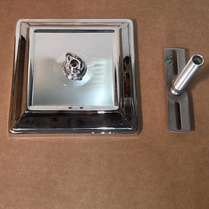 May include: A polished chrome shower head base, square in shape, with a central hook and a separate angled shower arm. The items are displayed on a brown surface.