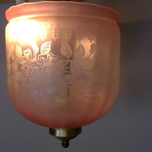 May include: A vintage-style, pink glass lampshade with a floral pattern. The etched design features flowers and leaves, and the shade is illuminated from within. The lampshade has a brass-colored base and a decorative finial.