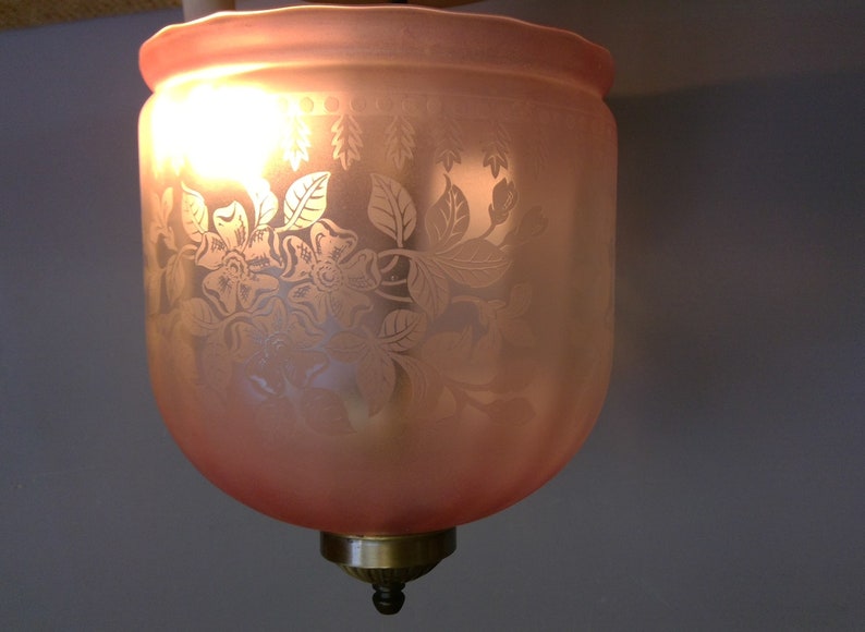 May include: A vintage-style, pink glass lampshade with a floral pattern. The etched design features flowers and leaves, and the shade is illuminated from within. The lampshade has a brass-colored base and a decorative finial.